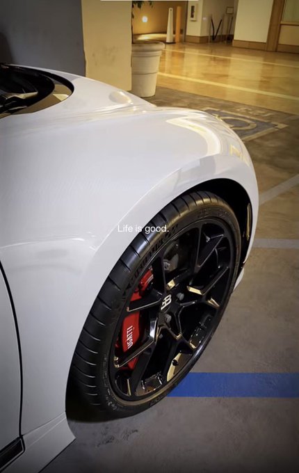 A close-up shot of a white sports car's front wheel, featuring black rims with red brake calipers, parked indoors with the caption 'Life is good.' overlaid.
