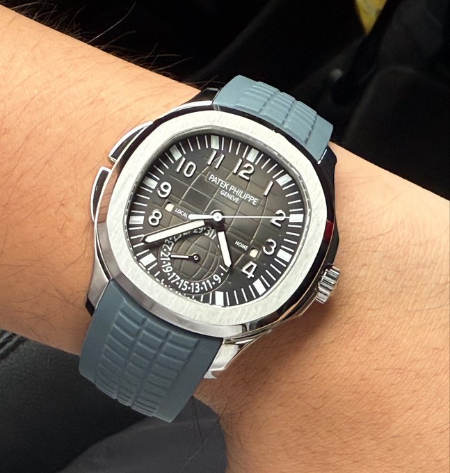 Close-up of a wrist wearing a Patek Philippe Aquanaut watch with a gray textured dial featuring local and home time complications, mounted on a light blue rubber strap.
