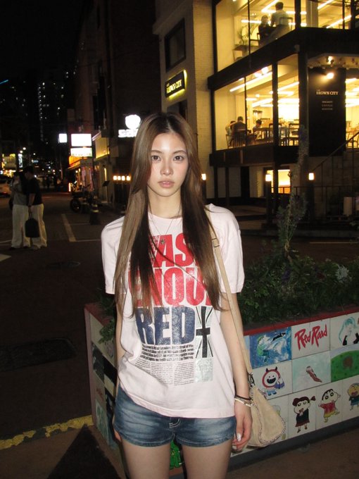 A young woman with long dark hair stands on a nighttime city street wearing a white graphic t-shirt with red and blue text and denim shorts.