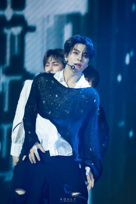 A K-pop idol performs on stage in a dark blue distressed sweater over a white collared shirt, with another performer visible behind him; the image is watermarked 'AQULY'.