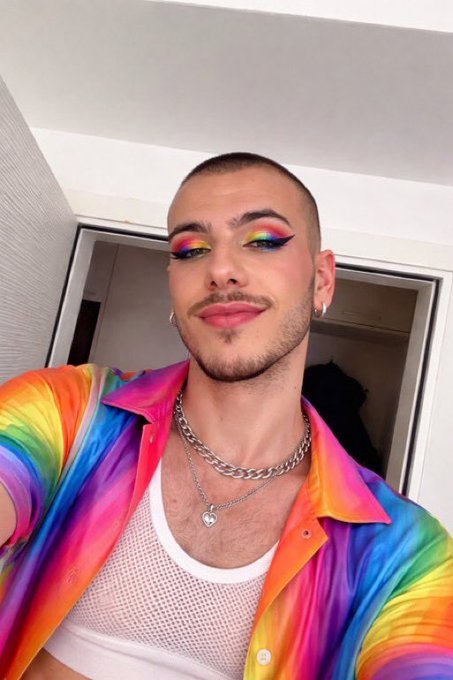 The image features a man with rainbow-colored hair, wearing a white shirt and a necklace that has a heart pendant. He is posing for a photo, looking directly at the camera while smiling. The background of the image shows a door or an open window, suggesting that he might be in a room or an area where people gather. The post caption reads "gm happy pride month y'all", which indicates that this could be related to Pride Month celebrations and is meant to express support for LGBTQ+ rights and diversity.