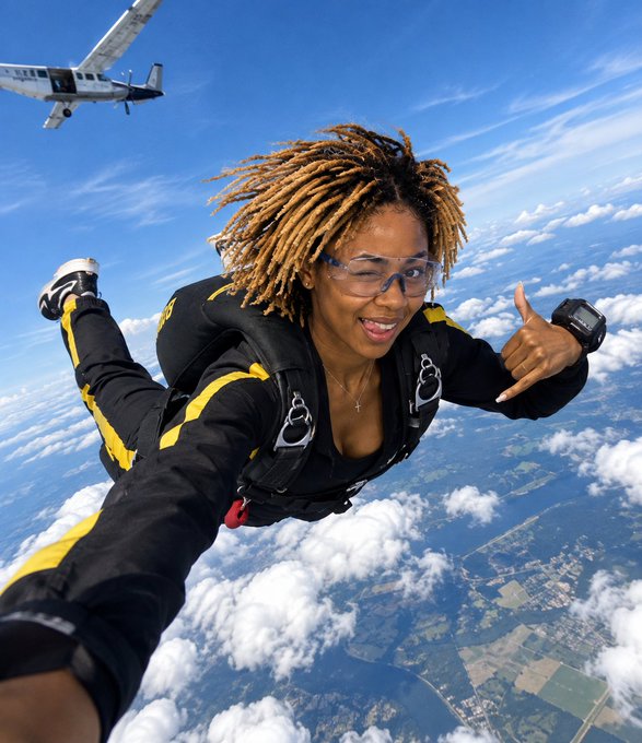 Action shot of a smiling woman with dreadlocks and glasses skydiving, making a shaka hand sign, with a small airplane visible in the upper left against a backdrop of clouds and land below.