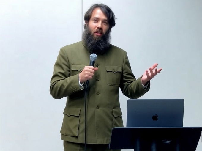 A bearded man in an olive-green military-style jacket stands at a podium with a microphone and an Apple laptop, gesturing mid-speech as he delivers a commentary on BIP110 and authoritarianism, his expressive pose suggesting a passionate critique of power dynamics within the political movement.