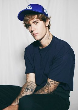 Justin Bieber, sporting a blue snapback cap, dark t-shirt, and visible tattoos, poses for a portrait. The image aligns with the post discussing his status as the favorite to headline the Super Bowl halftime show, as indicated by Polymarket traders.