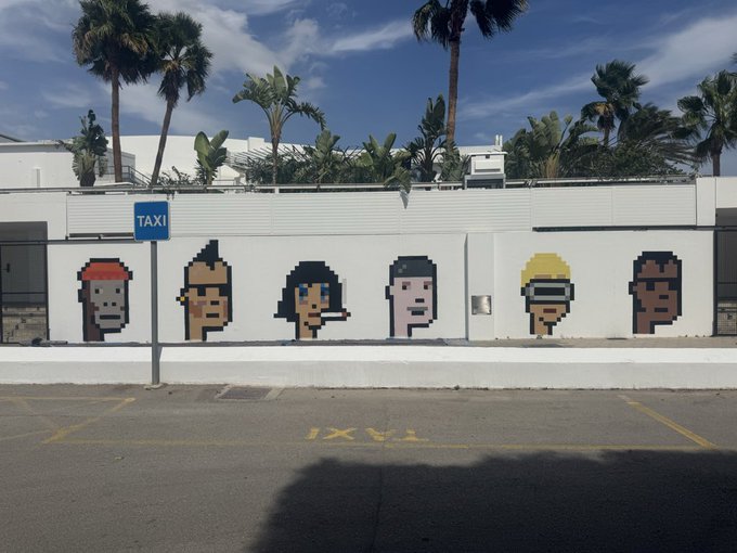 Pixel art murals of Gorillaz band members painted on a white wall at a taxi stand, with palm trees visible in the background against a blue sky.