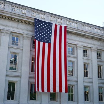 A massive American flag hangs vertically from the facade of a grand neoclassical government building, with its bold red and white stripes and blue field of stars prominently displayed against the light-colored stone exterior. The image visually underscores the post's message about US government efforts to reinforce the dollar's status as the world's reserve currency, symbolizing American financial authority and the nation's commitment to maintaining dollar dominance globally.