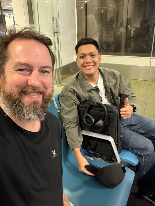 Two smiling men, one wearing a black t-shirt and the other giving a thumbs-up in a green jacket, sit in an airport waiting area with a black backpack and headphones between them. The image accompanies a post announcing Chad and Rondie (likely pictured) traveling to Bali to represent Sapien at Binance Coinfest, where they'll present "Proof of Quality."
