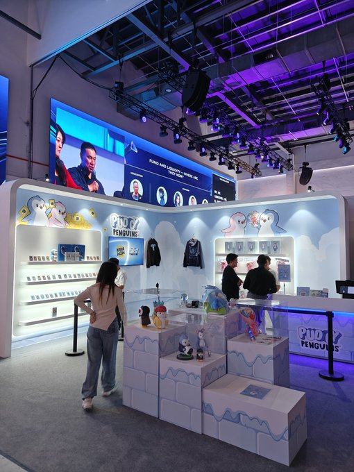 An interior view of a brightly lit 'Pudy Penguins' retail booth with merchandise displays, screens, and several customers browsing.