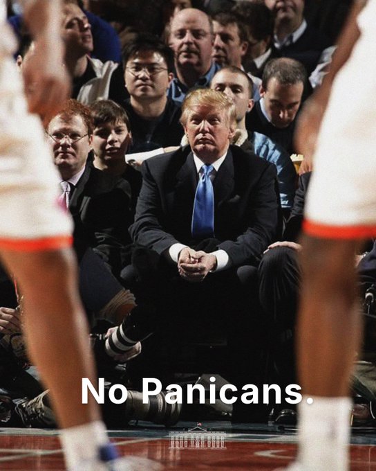 The image shows a basketball court with two men sitting down, one of whom appears to be wearing a suit and tie while the other has a baseball cap on his head. The background reveals that there are many people present, indicating a crowded event or gathering. A caption below the image reads "No Panics", suggesting a sense of calmness in the situation despite the crowd.