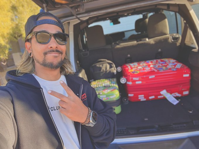 A young man in a backwards cap, sunglasses, and a hoodie throws up a peace sign in front of an SUV packed with colorful suitcases and a luggage tag, ready for a travel adventure—perfectly capturing the "Gm Fam" energy as he kicks off a trip with good vibes.