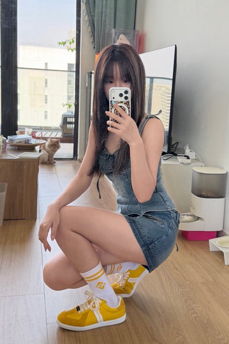 A young woman takes a mirror selfie while squatting on a wooden floor, wearing a denim outfit with yellow and white sneakers, with a cat visible in the background.
