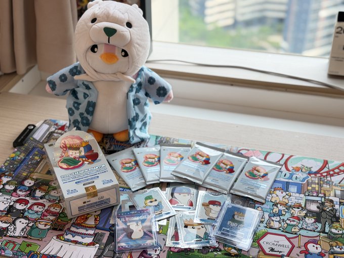 A photograph of a 'Cromwell' themed merchandise collection on a windowsill, featuring a plush polar bear wearing a hat, packaged pins, cards, and stickers spread across a patterned cloth.