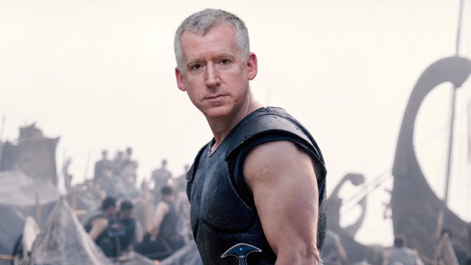 A man with short gray hair wearing black Roman-style armor stands in a battlefield setting with tents and soldiers behind him.
