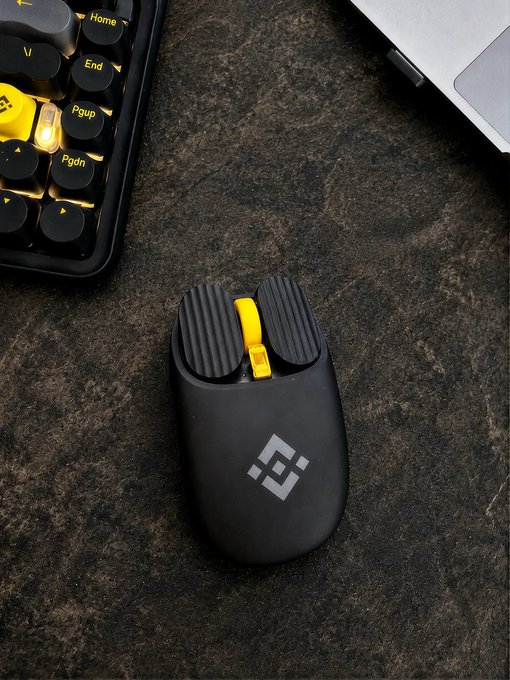 A black wireless mouse with a yellow scroll wheel and the Binance logo sits on a dark textured desk mat next to a keyboard with yellow-lit keys, fitting the "click to start the day" theme by showcasing the tools needed to begin cryptocurrency trading each morning.