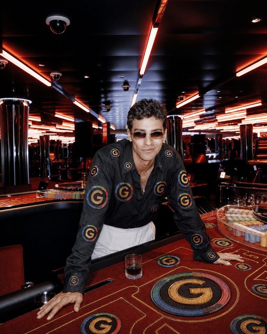 A man with dark hair and sunglasses leans on a gambling table covered in a red felt cloth with a repeating 'G' logo, set in a dimly lit, red-accented casino room.