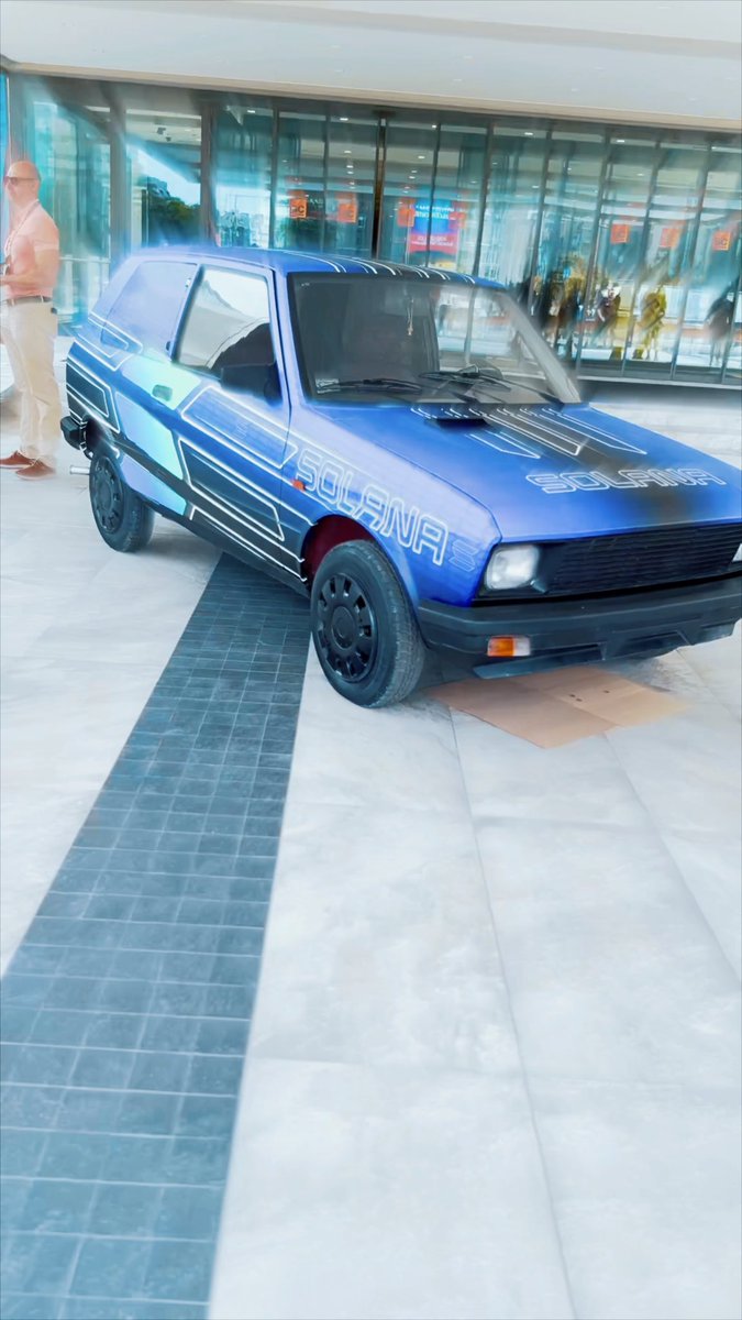 A blue compact car (appears to be a Tofaş/Fiat) parked in front of a glass-fronted building, wrapped with custom 'SOLANA' and 'SOLSURF' graphics and styling.