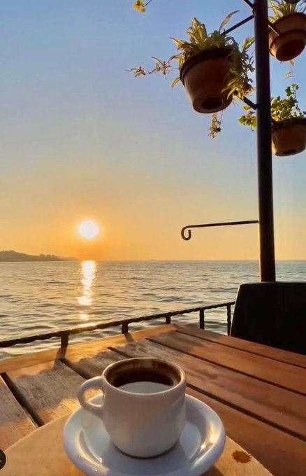 The image shows a cup of coffee sitting on top of a wooden table, with a view of the ocean and the sun rising behind it. The scene suggests that someone has just finished their morning coffee while enjoying the beautiful sunrise or sunset by the water.