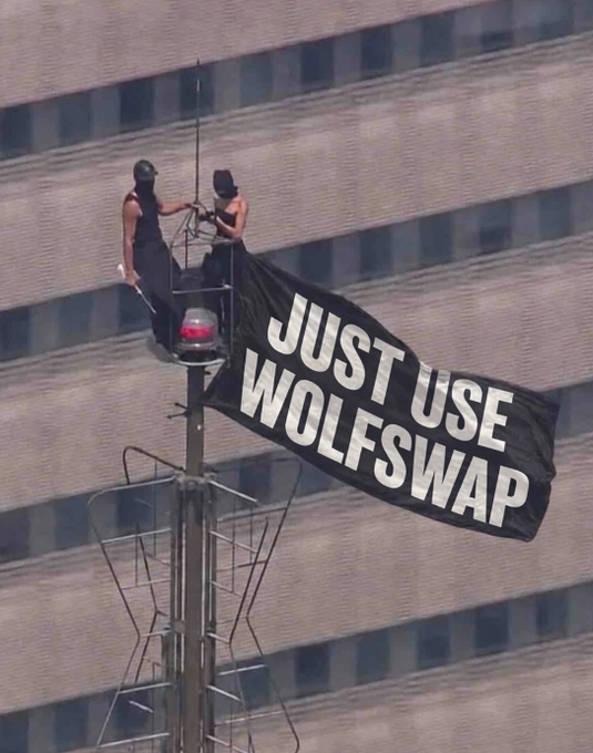 Two people on a tall structure holding a large black flag that reads 'JUST USE WOLFSWAP', with a building facade visible in the background.