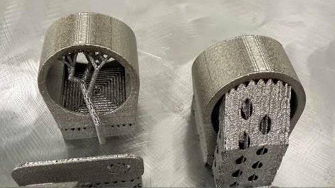 Two cylindrical metal 3D-printed parts with complex internal lattice structures rest on a textured surface, showcasing additive manufacturing capabilities.