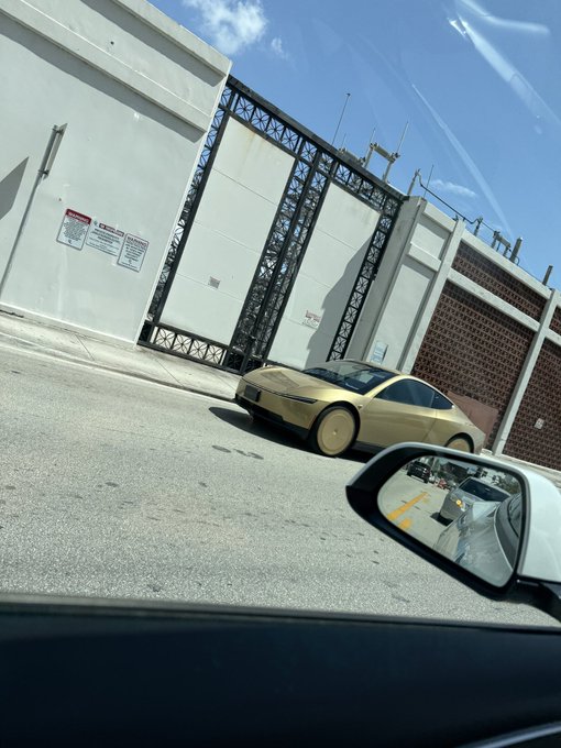 A gold-colored sports car, appearing to be a Tesla Cybertruck prototype, drives past a white industrial building, viewed through the side mirror of another vehicle.