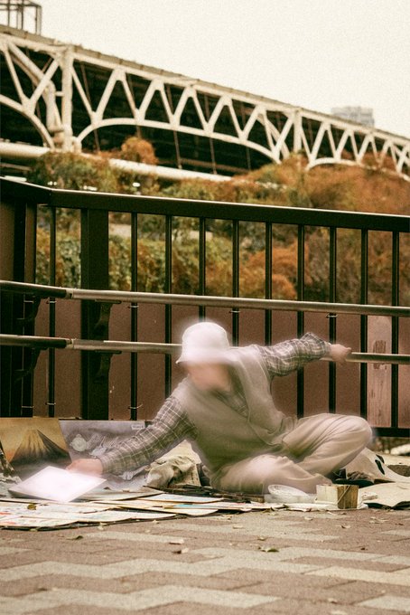 The image shows a man sitting on the ground with his arms outstretched, possibly as part of an art project or performance. The man appears to be wearing a hat and has papers around him, suggesting that he might be working on some creative work. The photo is taken from behind the man, giving it a sense of depth and perspective. The background features a bridge in the distance, adding an interesting element to the scene.