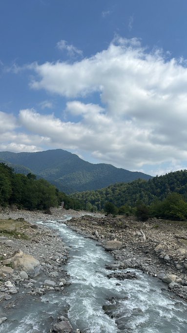 A scenic landscape photograph of a rushing river flowing through a rocky, mountainous valley under a partly cloudy blue sky.