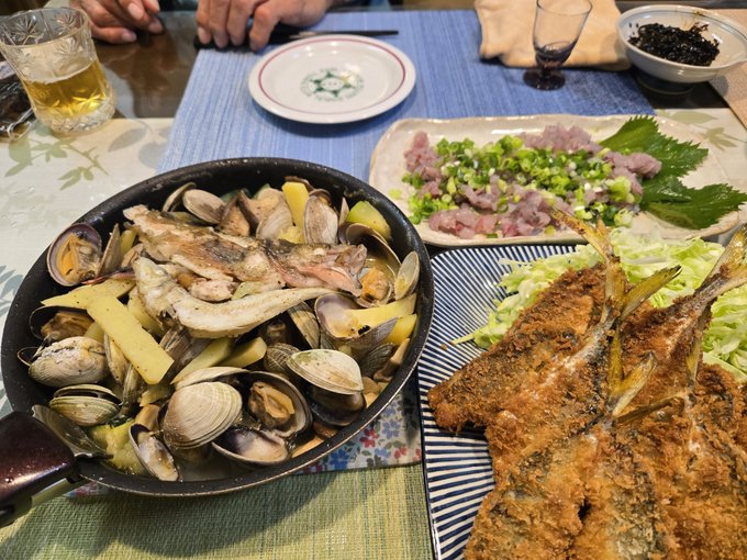 A meal spread featuring a pan of clams and grilled fish with vegetables, a plate of raw fish with green onions on shiso leaves, fried fish, and a beer glass on a blue table.