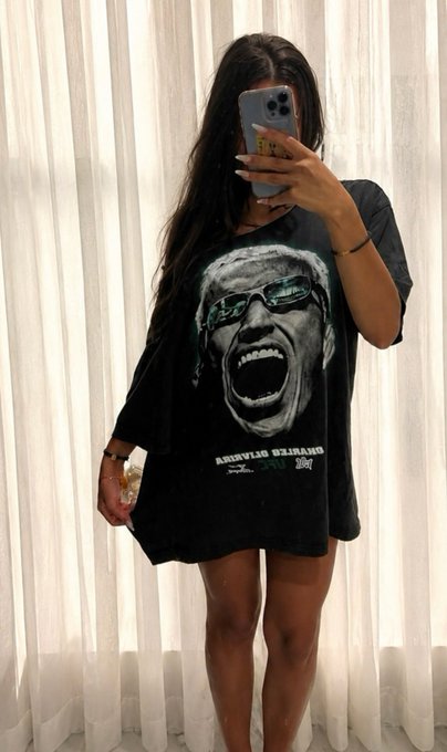 The image shows a woman taking a photo of herself with her mouth open, wearing a black t-shirt with a graphic design or poster that says "scotland has never beaten brazil. let's keep it that way fit for today's game" as part of her selfie. There is also an additional text in the post which starts with "@jup_predict". The woman is posing close to a curtain and there are multiple texts surrounding this scene, including "scotland has never beaten brazil", @jup_predict,"and let's keep it that way fit for today's game", as well as other descriptions of the image.