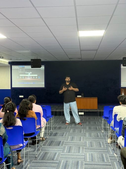 A photograph of a man standing in front of a projection screen displaying the Google homepage, giving a presentation to an audience seated in blue chairs in a conference room.