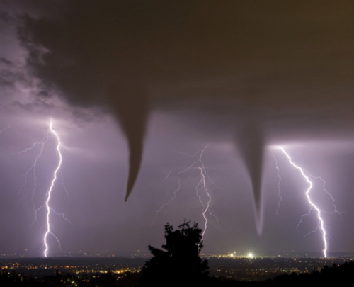 A dramatic nighttime photograph capturing two tornadoes illuminated by multiple lightning strikes over a city skyline.