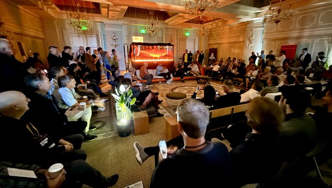 A large audience sits in an ornately decorated hall with chandeliers and patterned flooring, watching a presentation or panel discussion on a stage illuminated with red lighting.