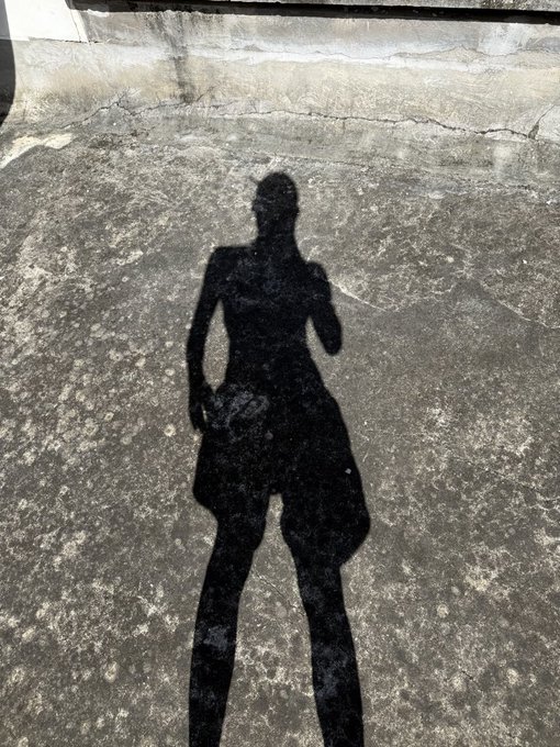 A shadow of a person wearing shorts is cast on a rough concrete pavement in bright sunlight, showing their silhouette standing upright.