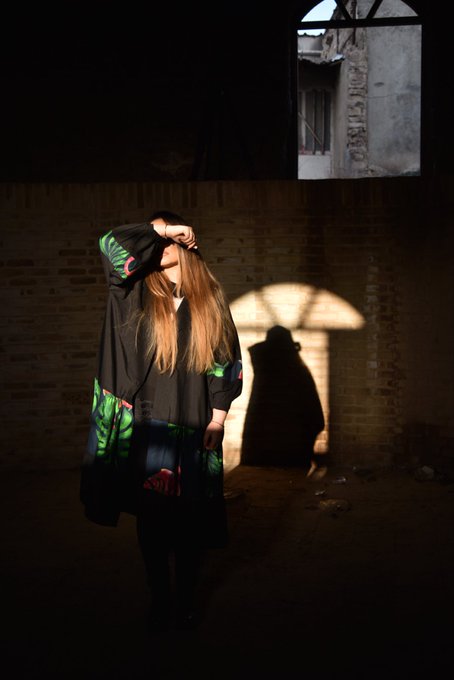 A moody, dimly lit photograph of a person with long hair wearing a dark jacket with green leaf designs, standing in a brick-walled space with sunlight casting a window-shaped shadow and a silhouette on the wall behind them.