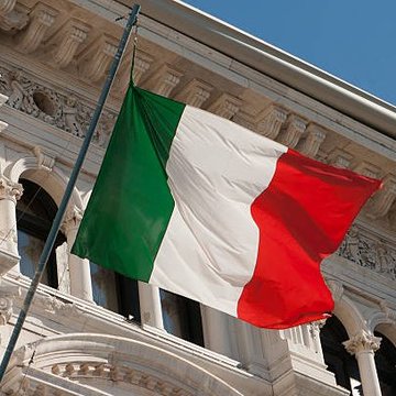 The image features a flag with green, white, and red colors flying from a pole attached to an old building. The flag appears to be waving in the wind, indicating that it's a sunny day. In the background of the image, there is a post with text written on it, which reads "LATEST- 34B€ @BancaSella becomes the first Italian bank to offer crypto related services #XRP and other #Altcoins". This suggests that the building might be an old Italian bank or financial institution. The flag flying from this building could symbolize the bank's commitment to innovation, embracing new technologies like cryptocurrencies, and staying ahead of the curve in their banking practices.