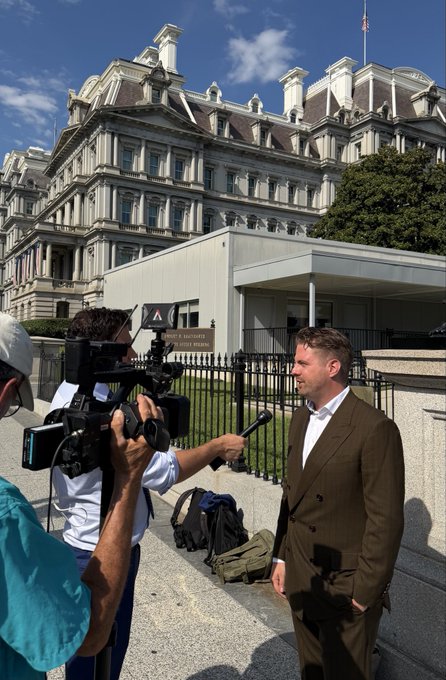 A man in a sharp brown suit is being interviewed outside the Eisenhower Executive Office Building, with a camera crew filming him as a reporter holds a microphone toward him. The scene captures a professional media moment, fitting the post's praise for his polished appearance as a representative of @blockchain and @askjuneai.