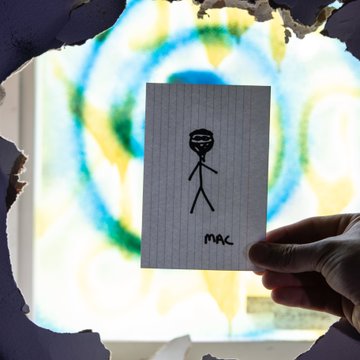 A hand holds a small piece of lined paper with a stick figure drawing labeled 'MAC' in front of a window decorated with a colorful circular mandala pattern.