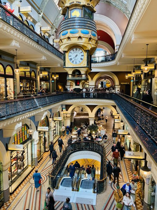 An interior view of an ornate multi-level shopping arcade featuring a large decorative clock, intricate iron railings, mosaic tile flooring, and visible storefronts including UGG and Mulberry.