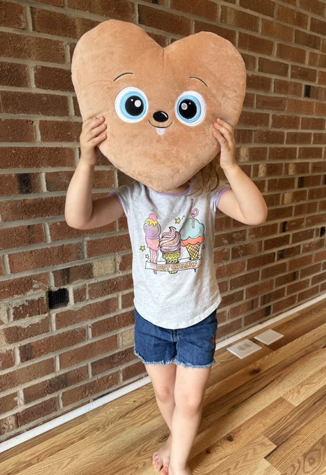 A young girl standing against a brick wall, wearing a gray ice cream graphic t-shirt and denim shorts, holding a large heart-shaped plush pillow in front of her face.