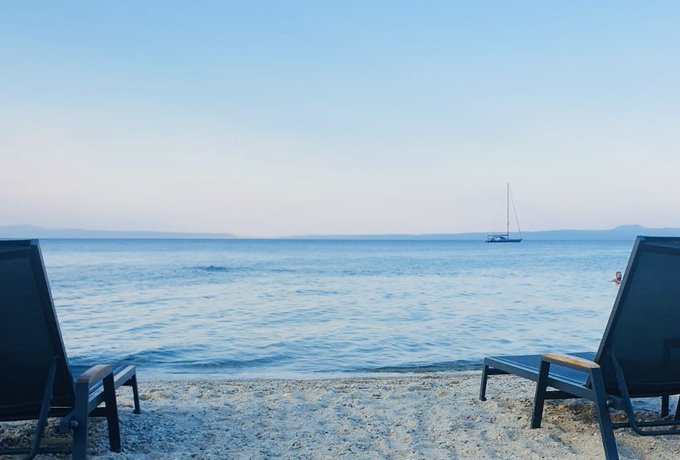 A serene beach scene at dusk with two empty dark lounge chairs in the foreground, calm blue water, and a sailboat anchored in the distance under a clear sky.
