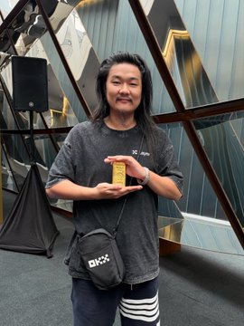 A woman with long dark hair wearing a gray t-shirt and a black crossbody bag stands in front of a modern geometric building, holding a small yellow object.