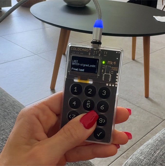 A close-up of a hand with red nail polish holding a 'COLDCARD Mk 4' hardware wallet device displaying 'LAST BATCH-signed P2bt Finalized' on its small screen.