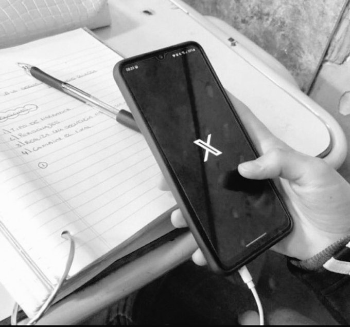 The image shows a person holding a black smartphone with an x symbol displayed on its screen, while sitting at a desk or table. In the background of the photo, there are some books visible, suggesting that this scene might be taking place in a study area or a workspace. The caption "gm say it back gmonad monad community" is likely a reference to a popular social media platform called Instagram, where users can share photos and engage with others by liking, commenting, and replying to posts.