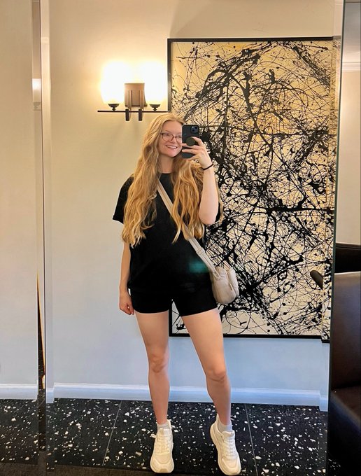 A young woman with long blonde hair and glasses takes a mirror selfie in what appears to be a restaurant or hotel lobby, wearing a black matching shorts set and white sneakers with a small crossbody bag. The large abstract black-and-white artwork behind her and her casual-yet-stylish outfit give off a polished, relaxed vibe perfect for a cheerful "Good morning friends" greeting.