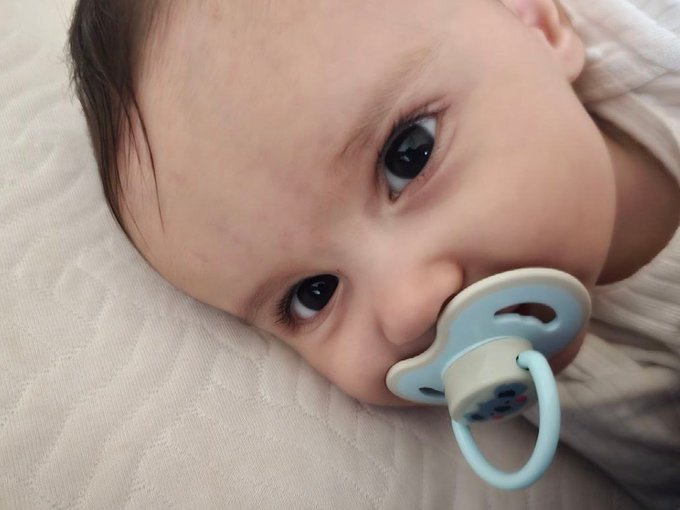 Close-up of a baby with a blue pacifier lying on a white textured blanket.