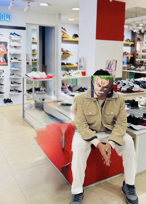 Person wearing a beige jacket and white pants sitting in a shoe store, with their face replaced by an anime character image (appears to be a One Piece character with green hair).