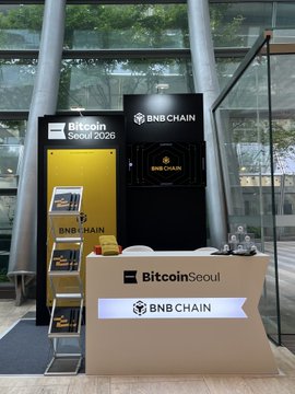 The image shows a large glass window with a sign that reads "Bitcoin Seoul 2016". In front of the window, there are two display cases showcasing various items related to Bitcoin. One case contains books about Bitcoin, while the other displays a TV screen showing Bitcoin logos and text. The overall setting appears to be an exhibition or event related to Bitcoin in Seoul.