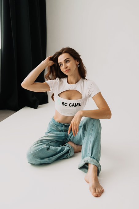 A young woman sits on a white studio floor wearing a white cutout crop top with 'BC.GAME' printed on it and baggy blue jeans.