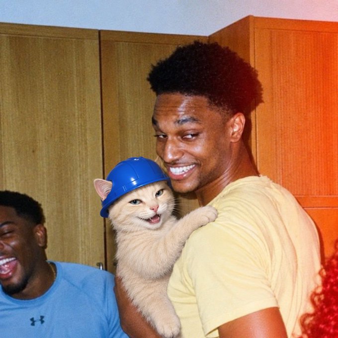 Basketball player Giannis Antetokounmpo smiles warmly while holding a cream-colored cat wearing a small blue hard hat.