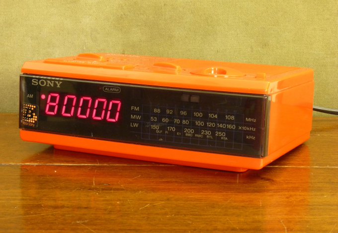 An orange vintage Sony alarm clock radio with red LED digits displaying '10:00:00', showing AM/FM/MW/LW frequency bands on a wooden surface.