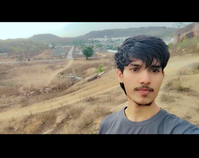 A selfie of a young man with dark hair and a light mustache and beard, wearing a grey t-shirt, with a dry, hilly landscape and a village in the background.
